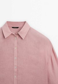 Chemise en lin rose clair à manches longues avec col pointu et boutons blancs sur le devant, présentée sur fond blanc.
