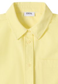 Chemise jaune boutonnée avec col et poche poitrine, comportant une étiquette blanche avec le texte "CECIL" à l'intérieur du col.