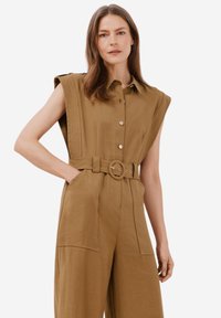 Ärmlös, brun jumpsuit med knappuppknäppning framtill, oversized axlar, bälte i midjan och stora sidofickor. Slät, texturerad tyg.