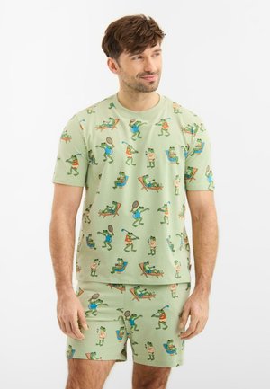Mies, joka pukeutunut vaaleanvihreään, samansävyiseen shortsien ja t-paidan settiin, jossa on piirroskuvia sammakoista pelaamassa golfia ja rentoutumassa aurinkotuoleissa.