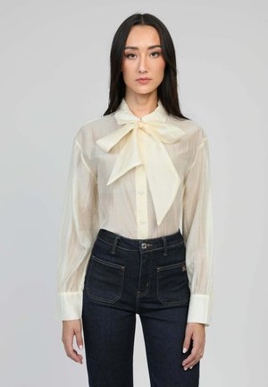 Blouse créme transparente avec un grand nœud au col, fermeture à boutons et manches longues ; associée à un jean taille haute bleu foncé.