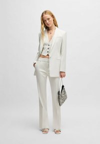 Weißer Anzug mit tailliertem Blazer und hochtaillierten Hosen, dazu eine verkürzte Weste. Strukturierte silberne Handtasche und Riemchensandalen.