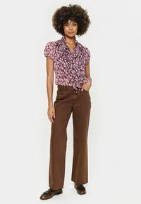 Rožinė trumpa rankovėmis gėlių bluzė su raukšlėtu apykaklė, derinama su rudomis plačiomis kelnėmis ir rudomis bateliais, stovinti ant neutralios fono.