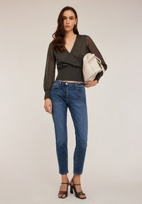 Top gri cu mâneci transparente striped, purtat cu blugi albaștri. Modelul ține o poșetă albă și poartă sandale negre cu toc.