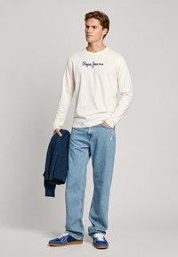 Langärmliges cremefarbenes Hemd mit dunkelblauer Schrift, hellblaue Jeans und blaue Sneakers; hält einen marineblauen gestrickten Pullover. Einfaches Design und lockere Passform.