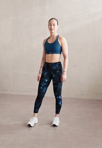 Sportininkė tamsiai mėlyno sportinio liemenėlė ir galaktikos raštų leggingai, vilkinti baltus sportinius batus; stovi ant lygaus, �šviesiai pilko grindų.