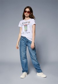 Camiseta gráfica blanca con el logo "GUESS", patrón azul y amarillo, combinada con jeans azules desgastados y zapatillas blancas. Modelo usando gafas de sol tintadas.