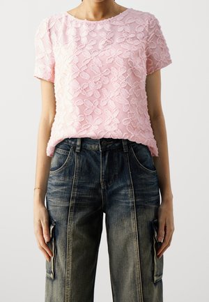 Femme portant un haut rose texturé à fleurs avec manches courtes, rentré dans un jean cargo en denim bleu foncé avec poches avant et latérales.