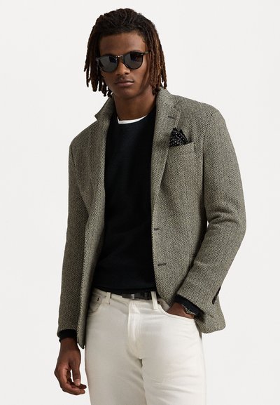 Blazer à motif pied-de-poule en gris et noir, associé à un pull sombre et un pantalon blanc, avec un mouchoir de poche et des lunettes de soleil.