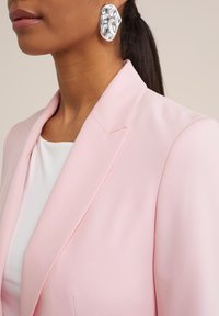 Helles rosa Blazer mit strukturierten Revers, kombiniert mit einem weißen Oberteil. Auffällige silberne abstrakte Ohrringe runden das Outfit ab.