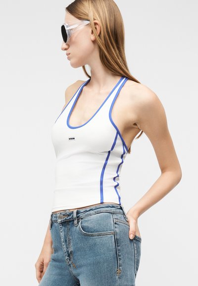 Γυναίκα που φοράει λευκό εφαρμοστό τοπ halter με μπλε περιθώριο και μπλε τζιν, στραμμένη προς τα αριστερά, με τα χέρια στις τσέπες και φοράει διαφανή γυαλιά ηλίου.