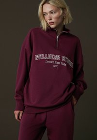 Bordowy cienki polar na zamek z białym napisem „WELLNESS STUDIO” i „Lower East Side NYC”. Miękki materiał, luźny krój, ściągacze przy rękawach.