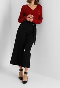 Top rojo de manga larga con escote en V, combinado con pantalones de tiro alto de pierna ancha negros con detalle de cinturón y tacones negros de punta.