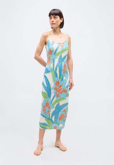 Γυναίκα που φοράει μια λευκή midi αμάνικη φόρεμα με μπλε, πράσινα και πορτοκαλί λουλουδάτα σχέδια, όρθια ξυπόλυτη μπροστά σε ένα απλό φόντο.