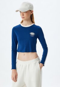 Blaues, kurz geschnittenes Langarmoberteil mit weißem Saum und "NEW YORK" Grafik. Kombiniert mit weißen Sweatpants mit elastischem Bund.
