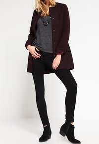 Burgunderfarbener Wollmantel mit rundem Ausschnitt, Frontknöpfen und gerippten Bündchen, kombiniert mit einem grauen Pullover und taillierten schwarzen Jeans. Ankle Boots runden den Look ab.