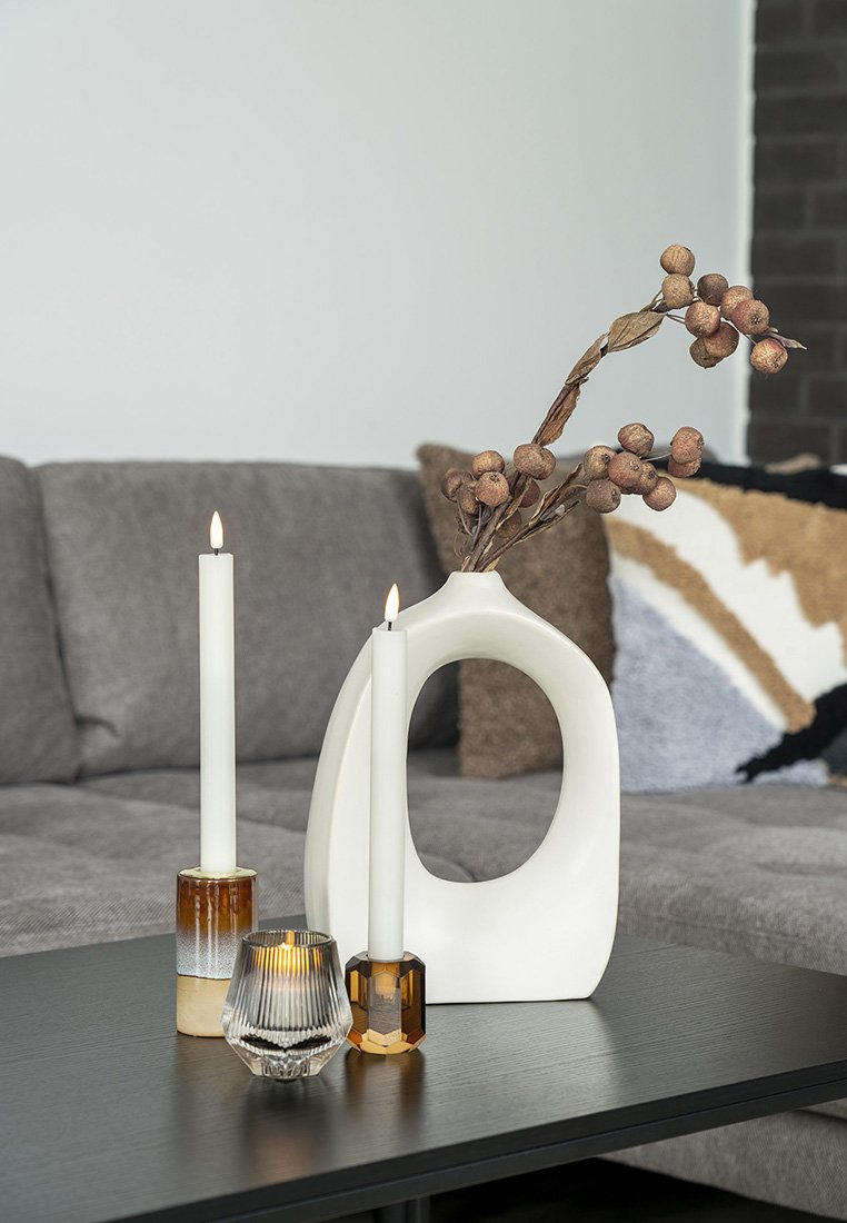 Vaso in ceramica bianca con un'apertura ovale, abbinato a due candele: una in vetro trasparente e una in vetro ambra, su un tavolo di legno scuro.