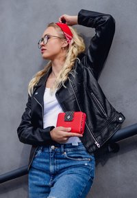 Black leather jacket, white shirt, blue jeans. Holding a red wallet with a patterned flap, against a gray background.
