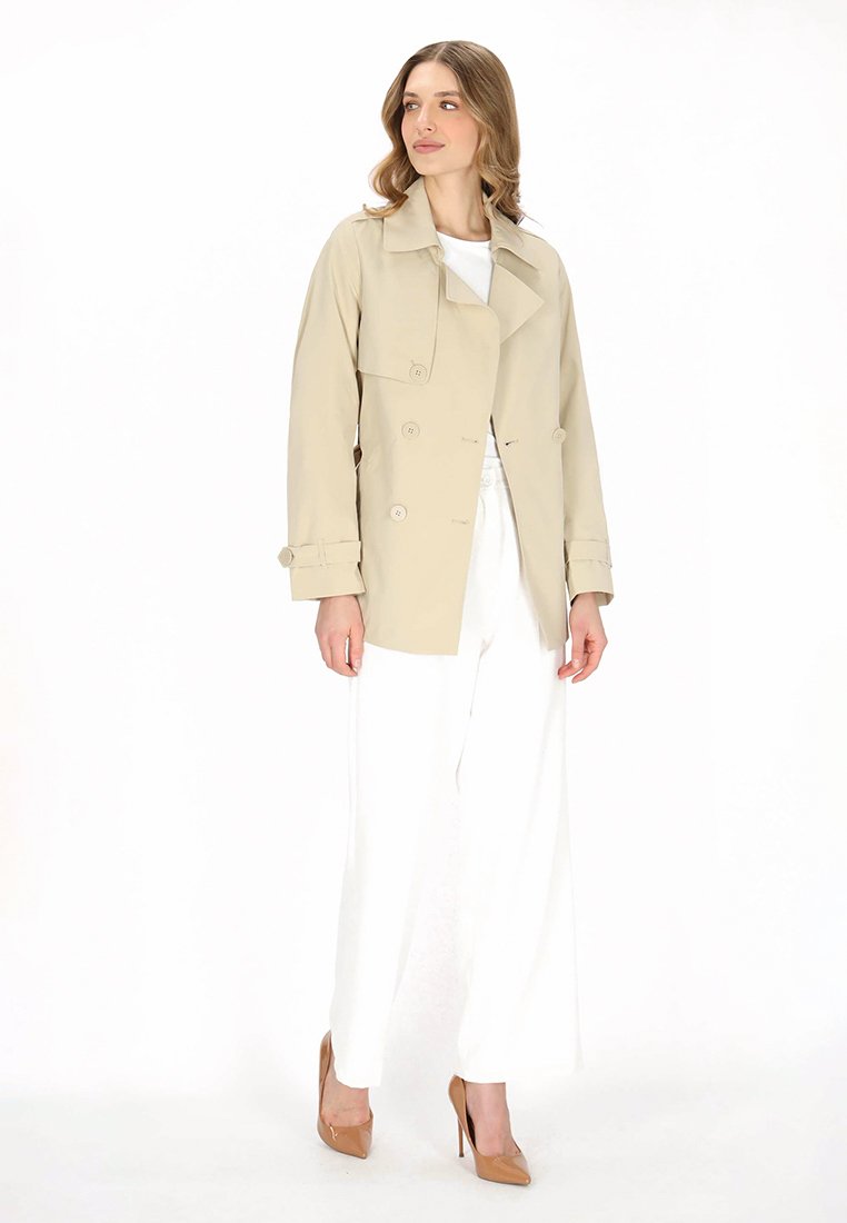 Beige trench coat with double-breasted design, button accents, and wide sleeves, paired with white wide-leg trousers and beige heels.