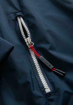 Navy-Jacke Detail: Nahaufnahme eines Reißverschlusses mit weißen Zähnen, der einen Metall-Zip-Puller mit ropem und einem schwarzen Plastikanhänger mit der Aufschrift "ombre" aufweist.