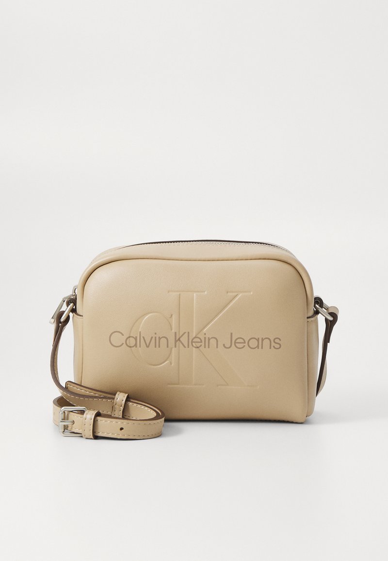 Borsa a tracolla in pelle beige con logo "CK" in rilievo, texture liscia, chiusura con zip e tracolla regolabile. Presenta un design minimalista.