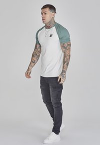 Tricou pentru bărbați cu un corp alb și manșete raglan verzi. Prezintă un mic logo negru pe piept. Echipat cu pantaloni cargo negri și adidași albi.