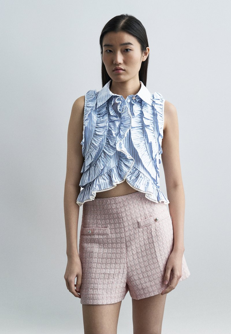 Νεαρή γυναίκα με μακριά σκούρα μαλλιά, φορώντας  αμάνικη μπλούζα με βολάν σε μπλε και λευκό και ροζ υφασμένα σορτς ψηλόμεσα, μπροστά σε απλό φόντο.