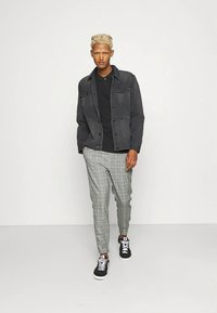 Čierna rifľová bunda cez čiernu polokošeľu, doplnená sivými károvanými nohavicami a čiernymi teniskami s bielymi podrážkami a šnúrkami.