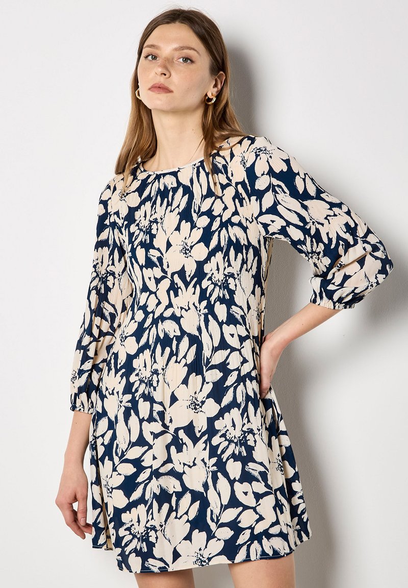 Blomstermønstret kjole i marineblå og creme. Har trekvartærmer, en lige pasform og en plisseret tekstur med diskrete detaljer.