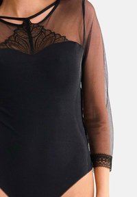 Bodysuit noir avec des manches longues en mesh transparent, un accent en dentelle au niveau du col, et une délicate bordure en dentelle au bas des manches.