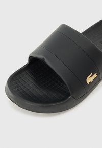 Svart sklidesandal laget av teksturert syntetisk materiale. Har en bred stropp med en subtil gulllogo og mønstret fotseng for grep.