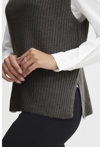 Ciemnoszara dzianinowa kamizelka z prążkowanym wzorem i bocznymi rozcięciami. Noszona na białej koszuli z długim rękawem. Gładka tekstura, luźny krój, warstwowy design.