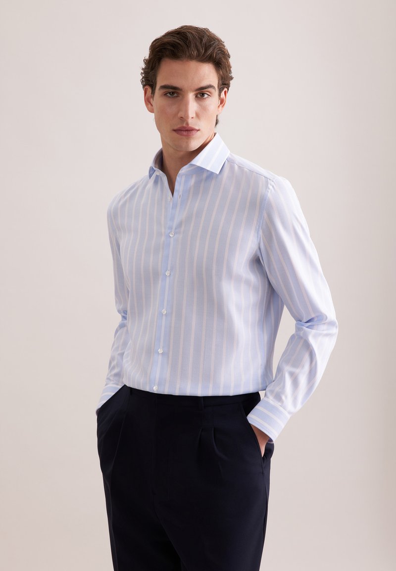 Homme portant une chemise à boutons bleu clair et blanc à rayures, un pantalon foncé, debout contre un fond clair uni, les mains dans les poches.