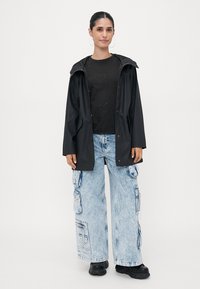 Black waterproof jacket with a hood, paired with a black top featuring heart studs and light blue cargo jeans. Black chunky shoes.