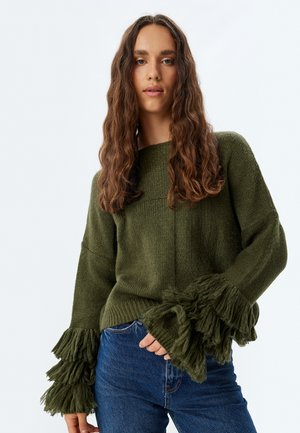 Green knitted sweater with a wide neckline and textured fringe details on the sleeves, paired with blue denim jeans.