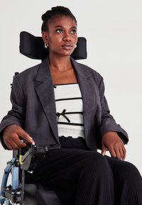 Γκρίζα σακάκι πάνω από λευκή υφή μπλούζα με μαύρες ρίγες, φ wears black ribbed παντελόνια. Το υποκείμενο κάθεται σε αναπηρικό καροτσάκι, χρησιμοποιώντας τον έλεγχο.