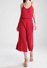 Mono rojo sin mangas y de corte holgado, con tejido texturizado, piernas anchas y recortadas, y una cinturilla ajustada. Combinado con sandalias beige con tiras.