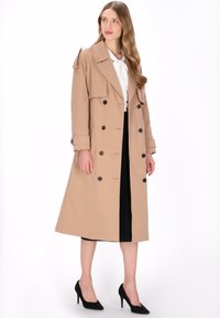 Beiger Trenchcoat mit doppelreihigem Design, großen Revers und Knopfverzierungen, ausgestattet mit einem taillierten Gürtel und knieumspielender Länge.