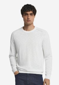 Weißer Rundhals-Pullover mit langen Ärmeln, strukturiertem Stoff und gerippten Bündchen. Wird über blauen Jeans getragen, Hände in den Taschen. Schlichtes Design.