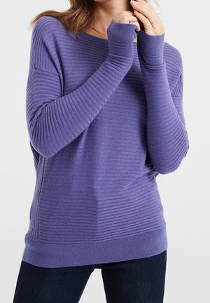 Femme portant un pull côtelé violet à manches longues avec des poignets allongés, debout devant un fond clair uni.