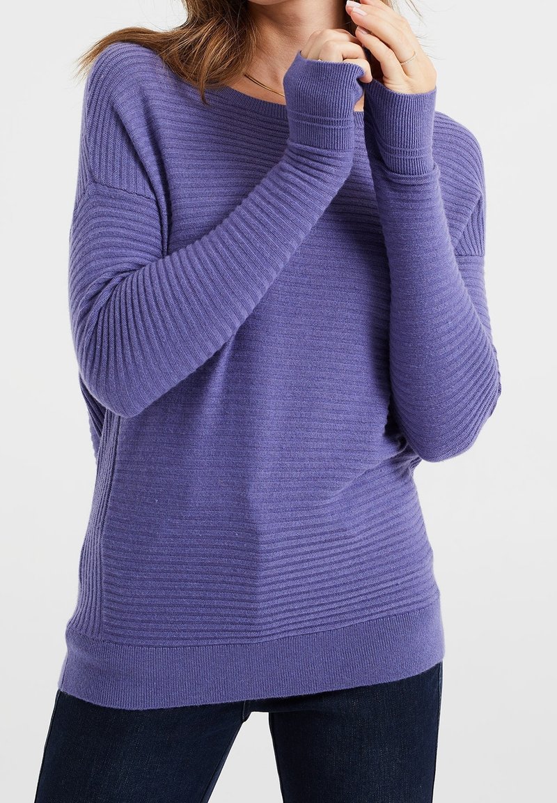 Femme portant un pull côtelé violet à manches longues avec des poignets allongés, debout devant un fond clair uni.