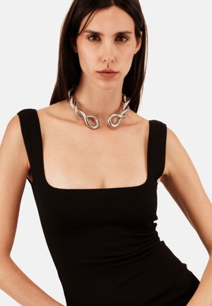 Frau mit langen dunklen Haaren, die ein schwarzes ärmelloses Kleid mit quadratischem Ausschnitt und eine breite silberne gedrehte Halskette trägt, vor einem schlichten Hintergrund.