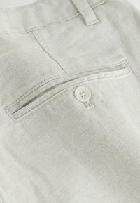 Poche de pantalon en tissu beige clair avec fermeture par bouton et passants de ceinture au-dessus, montrant des coutures détaillées et la texture.
