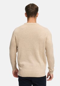 Jersey beige con mangas largas, cuello redondo y puños acanalados. Textura suave con ligeras variaciones en la tela.
