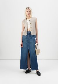 Beige strikket ermeløs vest med knapper, hvit crop topp, lys blå denim maxiskjørt med frontsplitt, og svarte sko. Holder en teksturert veske.