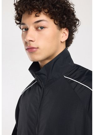 Jonge man met krullend haar draagt een zwart jack met ritssluiting en witte bies, kijkt iets opzij tegen een lichte achtergrond.