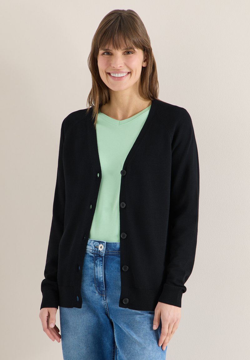 Femme souriante aux cheveux bruns portant un cardigan noir boutonné sur une chemise vert clair et un jean bleu, sur un fond uni.