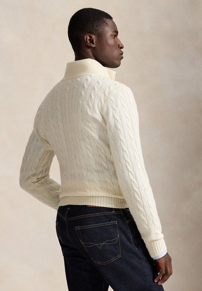 Uomo con maglione lavorato a trecce di colore crema e jeans blu scuro, di profilo su sfondo neutro.