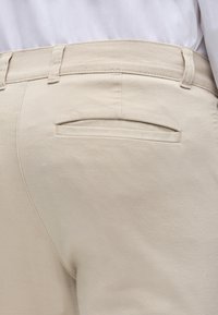 Κοντινό πλάνο σε μπεζ παντελόνι με πίσω τσέπη τύπου welt και θηλιές για ζώνη, φορεμένο με λευκό πουκάμισο.