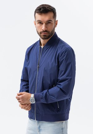 Dunkelblaues Bomberjackett mit Reißverschluss, gerippten Bündchen und Saum, seitlichen Taschen und einer glatten Stoffoberfläche. Kombiniert mit hellblauen Jeans.